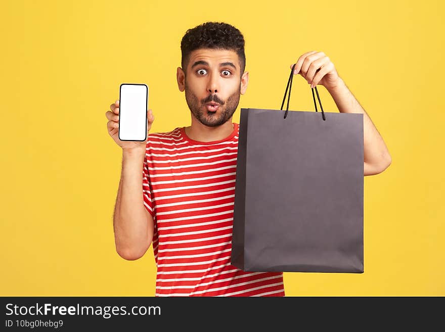 Surprised bearded man in red striped t-shirt holding in hands smartphone with white display and shopping bag, shocked with easy shopping online. Indoor studio shot isolated on yellow background