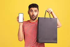 Surprised Bearded Man In Red Striped T-shirt Holding In Hands Smartphone With White Display And Shopping Bag, Shocked With Easy Stock Photos