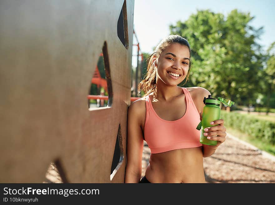 Attractive sportive young mixed race girl smiling at camera and holding water bottle while taking a break after working