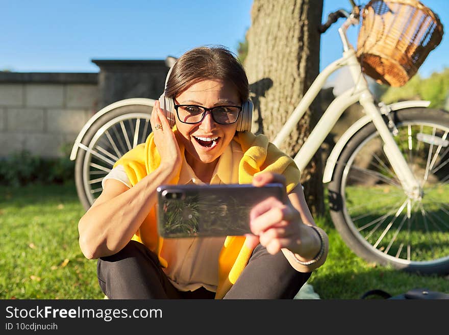 Mature woman in headphones with smartphone looking in screen of gadget, video, call. Happy female sitting on green lawn in park on sunny summer day. Mature woman in headphones with smartphone looking in screen of gadget, video, call. Happy female sitting on green lawn in park on sunny summer day