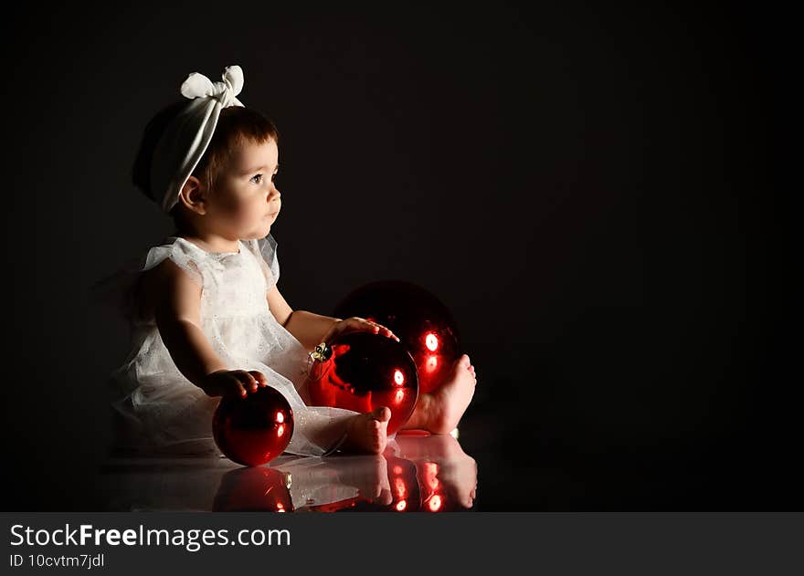 Little female in white headband and dress, barefoot. She holding two red balls, sitting on floor. Twilight, black background.