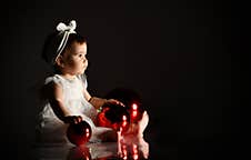 Little Female In White Headband And Dress, Barefoot. She Holding Two Red Balls, Sitting On Floor. Twilight, Black Background. Royalty Free Stock Photos