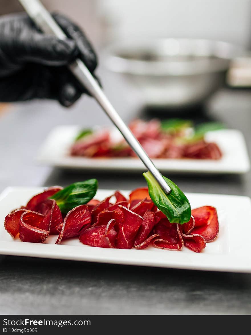 Chef placing basil leaves on sliced prosciutto plate with tongs