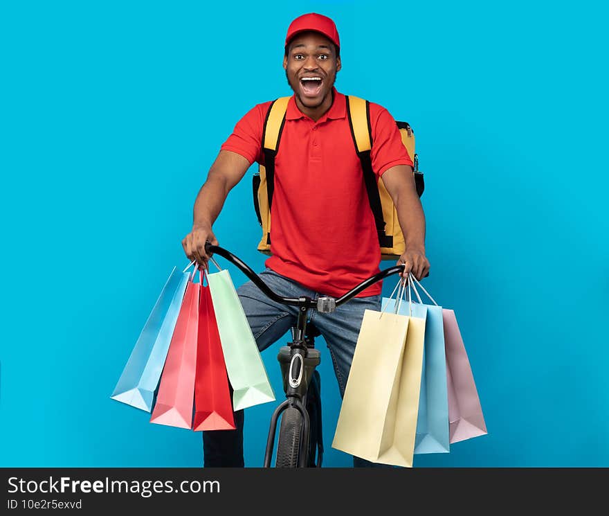 Delivery Service. Funny african american courier in red uniform and cap holding colorful shopping bags with customer`s order while riding a bicycle with wide open mouth, blue studio background. Delivery Service. Funny african american courier in red uniform and cap holding colorful shopping bags with customer`s order while riding a bicycle with wide open mouth, blue studio background