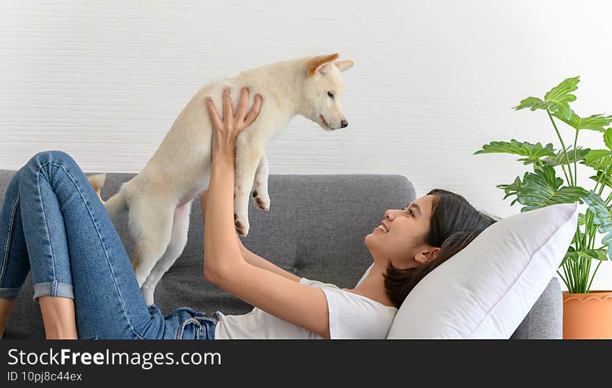 Beautiful young asian woman lying on sofa and playing with white Shiba Inu puppy in living room at home.