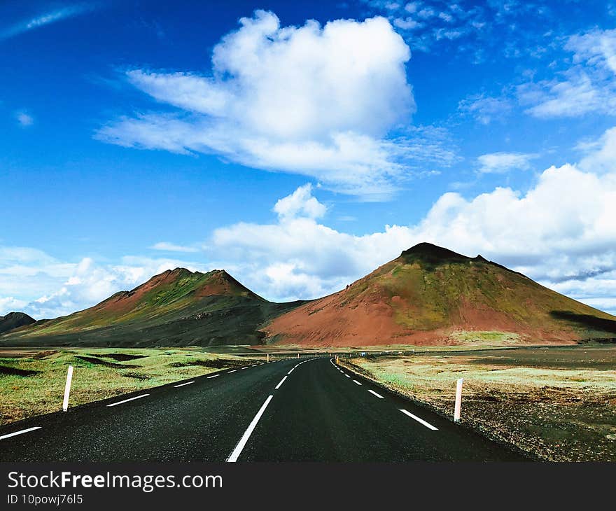 Road in a field surrounded by hills under a blue cloudy sky and sunlight