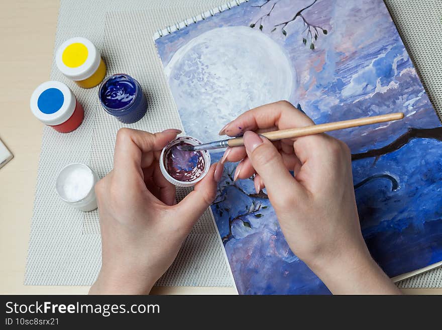 A close-up of female hands over a painted picture holding a brush and a cover for watercolors on which the colors are mixed next