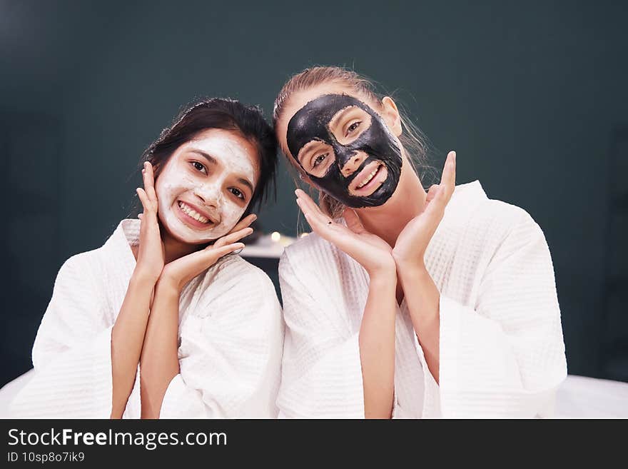 Caucasian and Asian woman in white bathrobe and black facial mask on face for treatment skin care together at spa. Lifestyle and