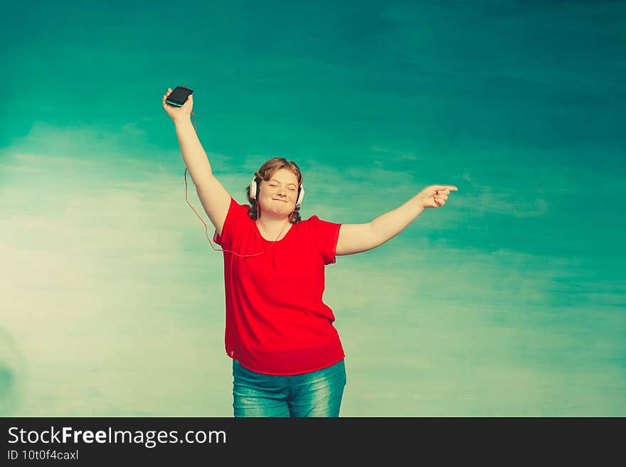 Young attractive plump woman with red hair and jeans jumping and fooling around and dancing in headphones on a blue background