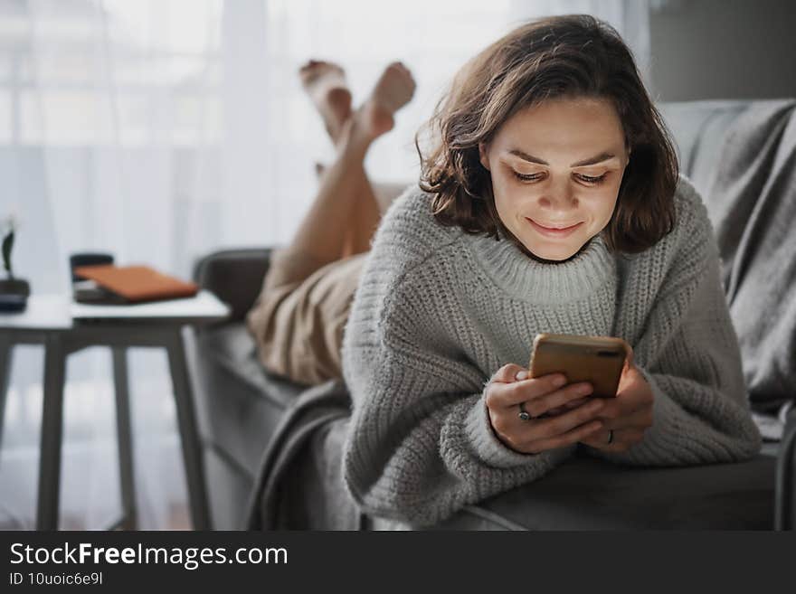 Happy young curly woman in gray sweater lying on sofa video chatting in mobile phone