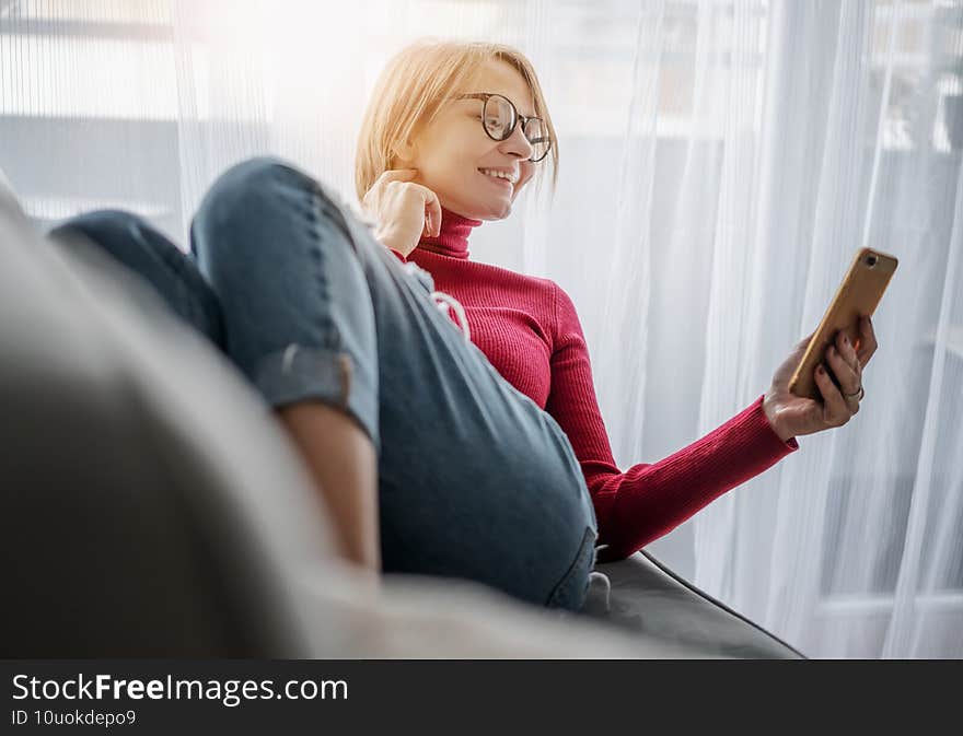 Beautiful smiling happy woman in a red turtleneck at home on a gray sofa lying with a mobile phone video chatting