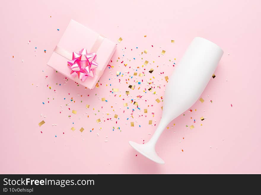 White champagne glass and gift box with confetti on pink background. Minimal party concept