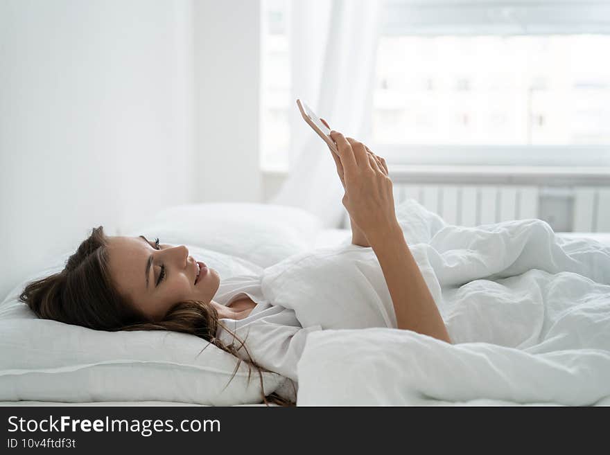 Smiling young woman using mobile smart phone, lying and relaxing in white comfortable bed at home