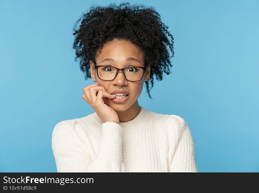 Close up studio portrait of shy awkward young Afro American woman wear glasses white sweater, biting nails feeling embarrassed, confused and nervous, looking at camera, isolated on blue background. Close up studio portrait of shy awkward young Afro American woman wear glasses white sweater, biting nails feeling embarrassed, confused and nervous, looking at camera, isolated on blue background