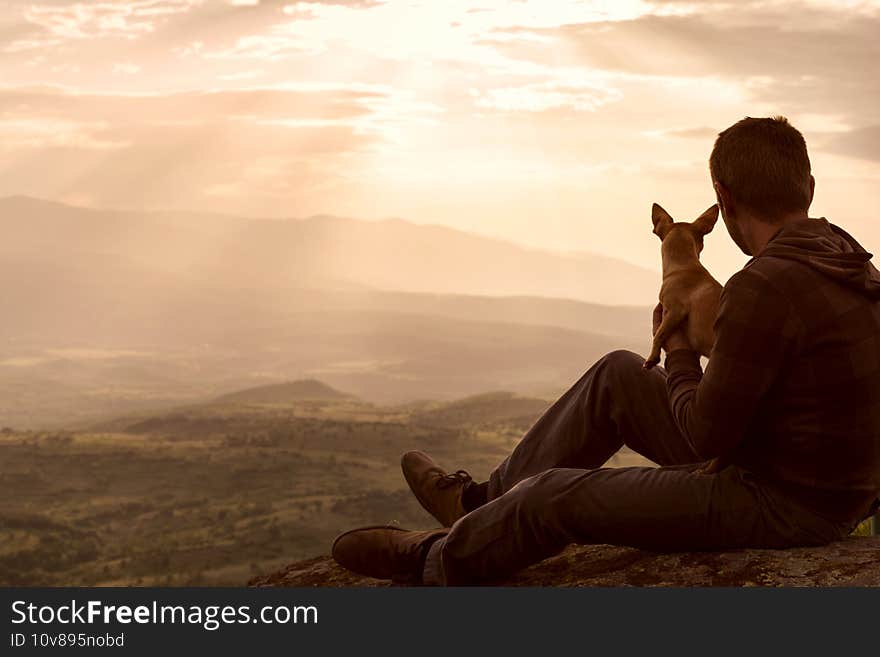 Young man spending time together with his dog pet, retiring in nature in top of hill and watching sunset in Rhodope Mountains, Bulgaria. Solitude, local tourism, social distancing concept. Back view. Young man spending time together with his dog pet, retiring in nature in top of hill and watching sunset in Rhodope Mountains, Bulgaria. Solitude, local tourism, social distancing concept. Back view