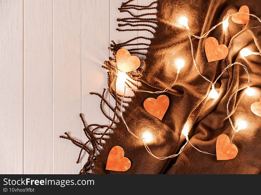 Paper hearts and a glowing garland are on a brown fringed scarf on a light wooden background.