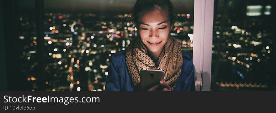 Woman texting instant messaging text on mobile phone online at night - bright light from screen illuminating young Asian girl in London city background banner panorama.