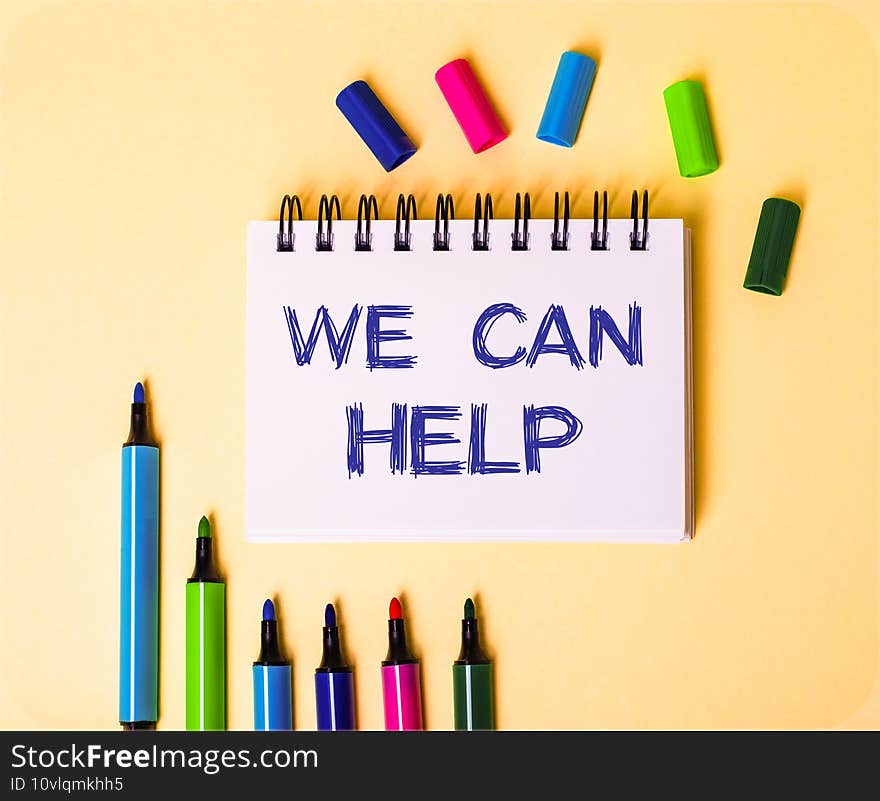 The words WE CAN HELP written in a white notebook on a beige background near multi-colored markers..