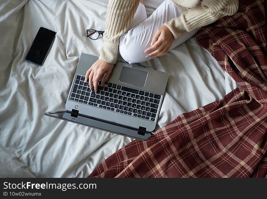 Young beautiful girl in cozy sweater in bed at home working on a laptop