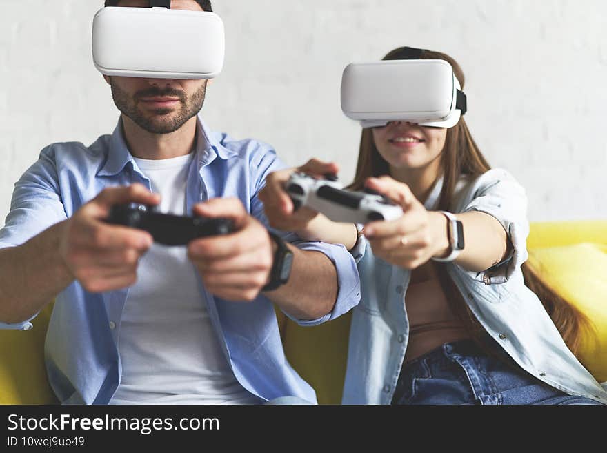 Closeup view of young smiling couple in VR glasses playing video game on yellow couch while having virtual reality experience. Closeup view of young smiling couple in VR glasses playing video game on yellow couch while having virtual reality experience