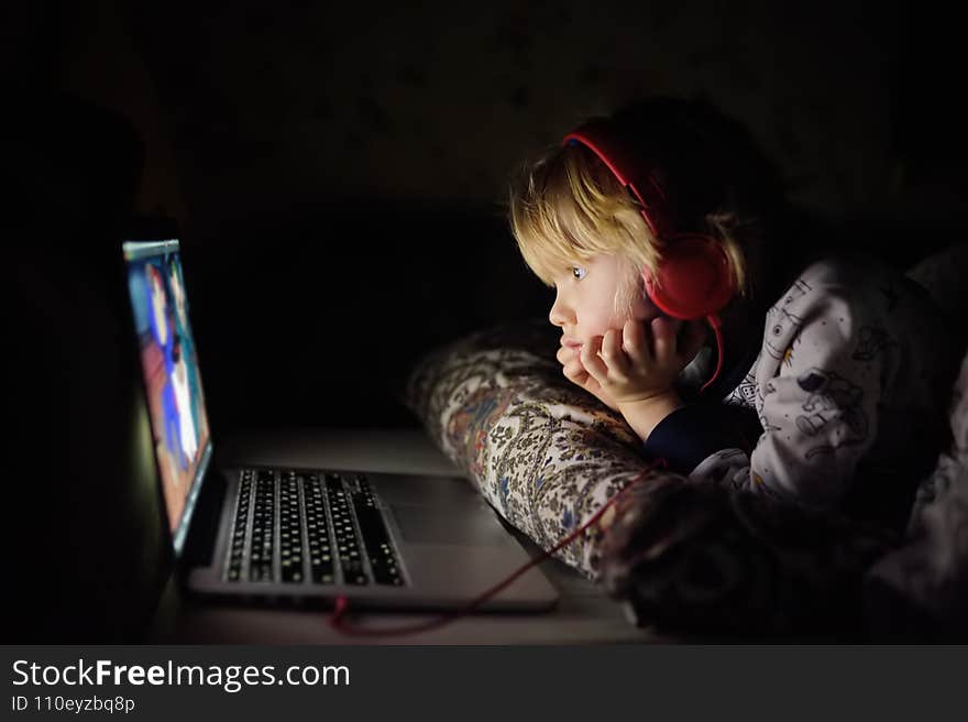 Cute preschool boy wearing headphones is watching cartoon movie by laptop before sleeping in bed or early in the morning right