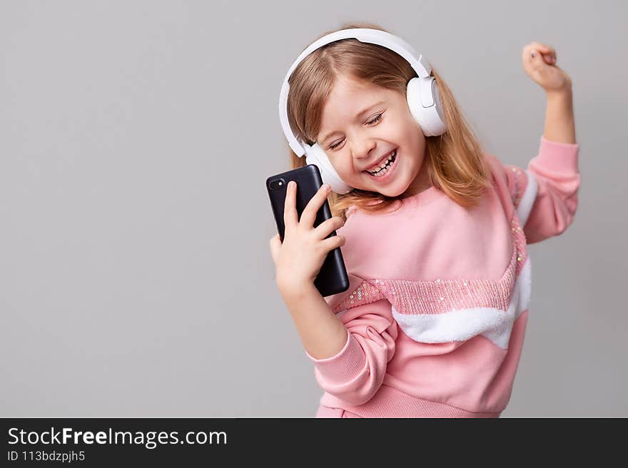 A little girl blogger influencer records a blog video on a smartphone, dances and listens to music with headphones on a