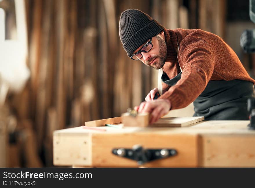 Focused adult male woodworker in apron and knitted gat cutting wooden plank while working in carpentry workshop. Focused adult male woodworker in apron and knitted gat cutting wooden plank while working in carpentry workshop