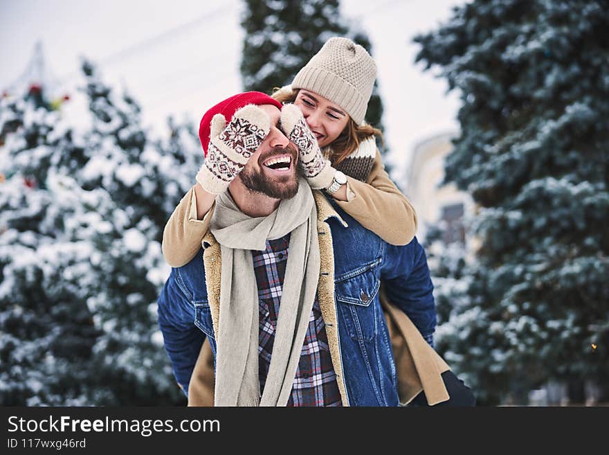 Smiling women is closing eyes of jolly boyfriend while riding him in forest among snow. Smiling women is closing eyes of jolly boyfriend while riding him in forest among snow
