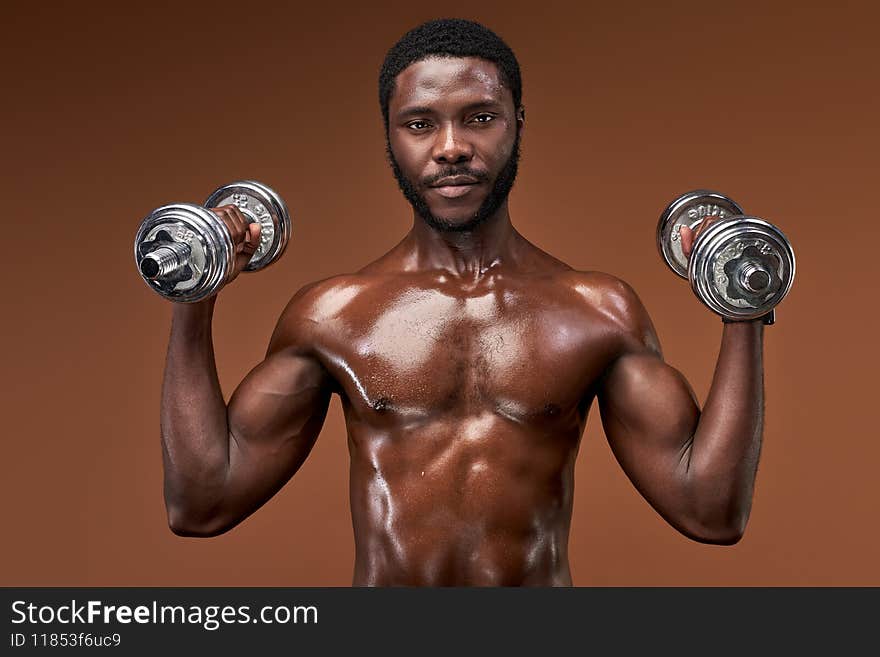 african bodybuilder guy doing exercises with dumbbells in studio on brown background, naked muscular male posing. african bodybuilder guy doing exercises with dumbbells in studio on brown background, naked muscular male posing