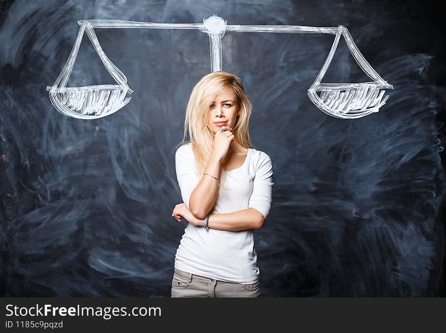 Beautiful woman is thinking, weighing the situation, making a choice in the studio near a chalk board with drawn scales. Creative