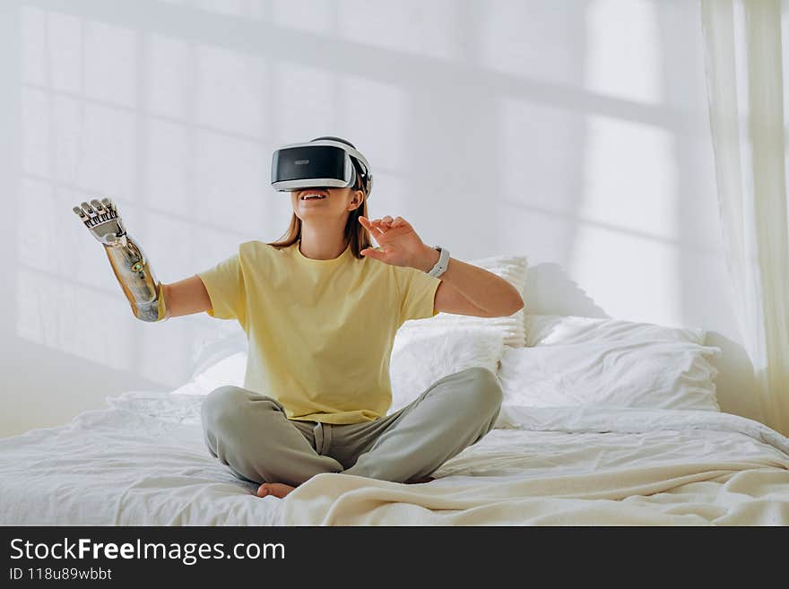 Happy woman with a modern robotic bionic prosthetic arm wearing virtual reality glasses sits on a bed in comfortable home clothes on a sunny day in a bright room, raising her hands and smiling. Touches the air using modern virtual reality glasses. Happy woman with a modern robotic bionic prosthetic arm wearing virtual reality glasses sits on a bed in comfortable home clothes on a sunny day in a bright room, raising her hands and smiling. Touches the air using modern virtual reality glasses.