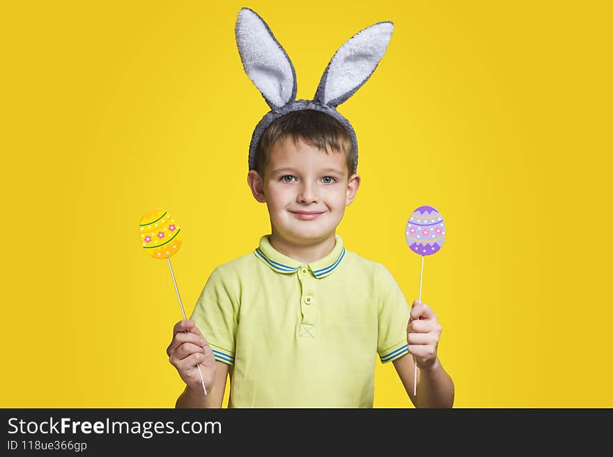 Cute boy with bunny ears holds colored eggs in his hands on yellow background. Happy child boy with easter eggs and bunny ears on yellow background.