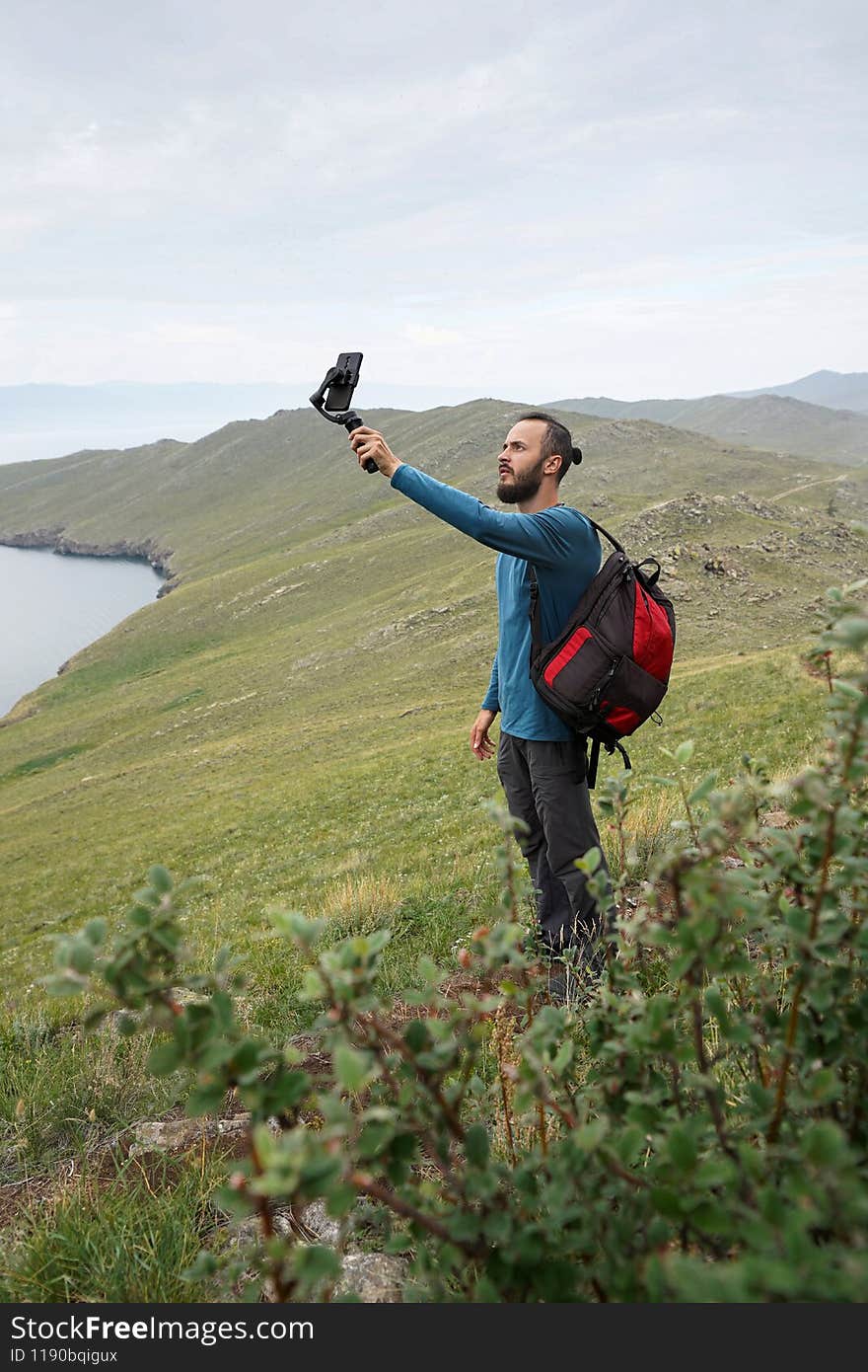 Tourist man with backpack takes a selfie on the shore of the bay Travel photography Grey sky Green hills. Tourist man with backpack takes a selfie on the shore of the bay Travel photography Grey sky Green hills