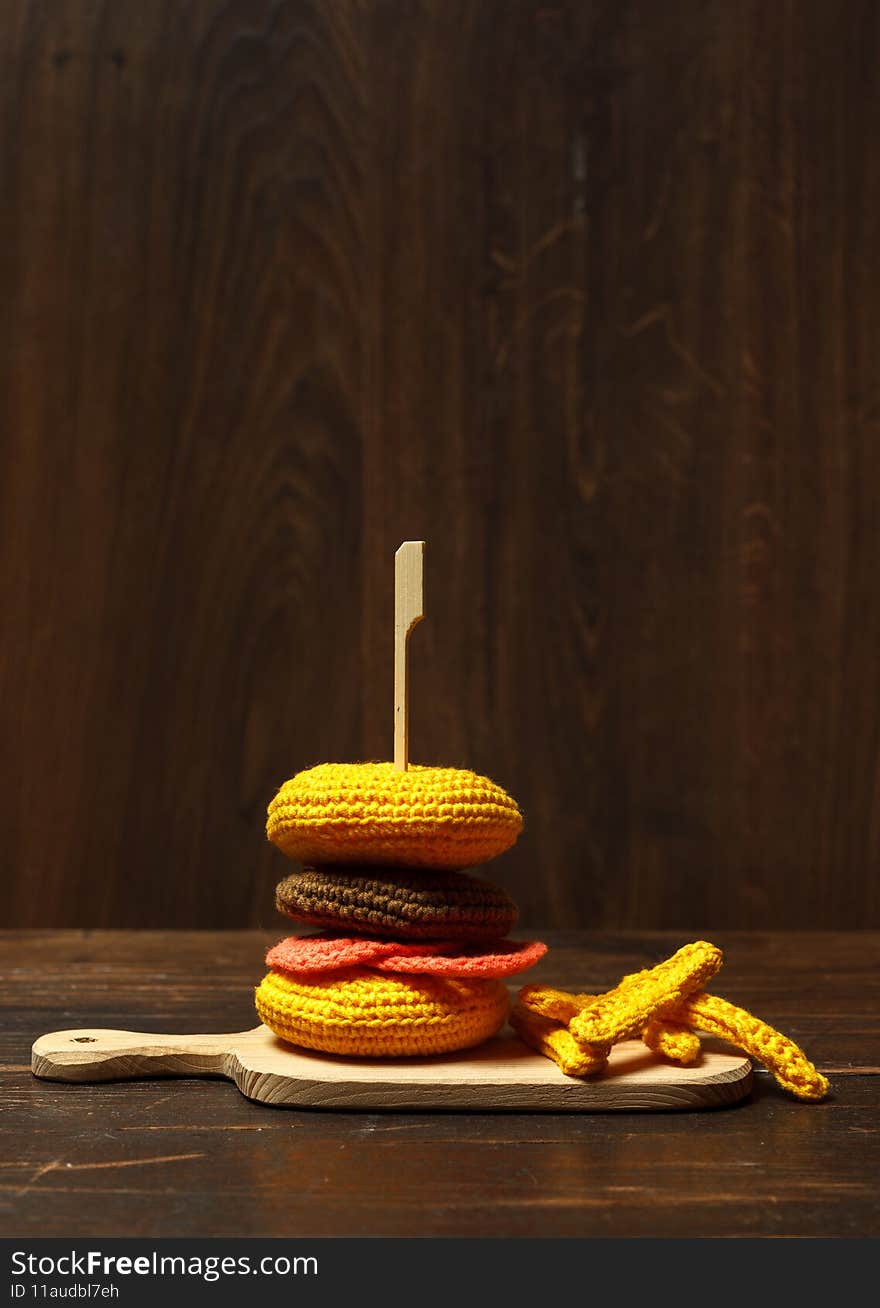 Knitted food. Fast food. Toy hamburger amigurumi and French fries on a wooden board. Close-up on a wooden background, low key, vertically with space. Knitted food. Fast food. Toy hamburger amigurumi and French fries on a wooden board. Close-up on a wooden background, low key, vertically with space