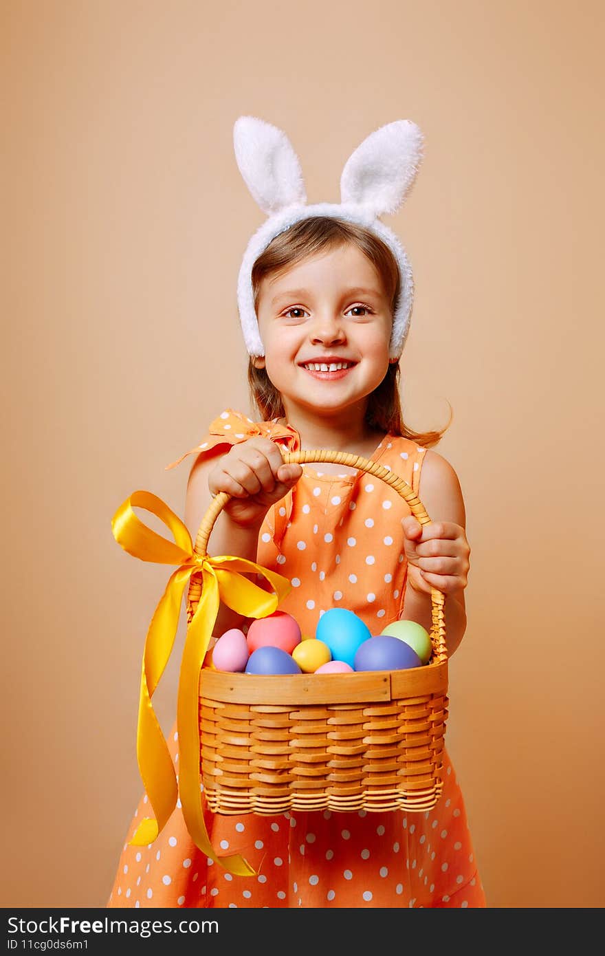 A cheerful girl with rabbit ears on her head and a protective mask with a basket of colored eggs in her hands on a beige background. Funny crazy happy baby. Covid Easter to the child. A cheerful girl with rabbit ears on her head and a protective mask with a basket of colored eggs in her hands on a beige background. Funny crazy happy baby. Covid Easter to the child