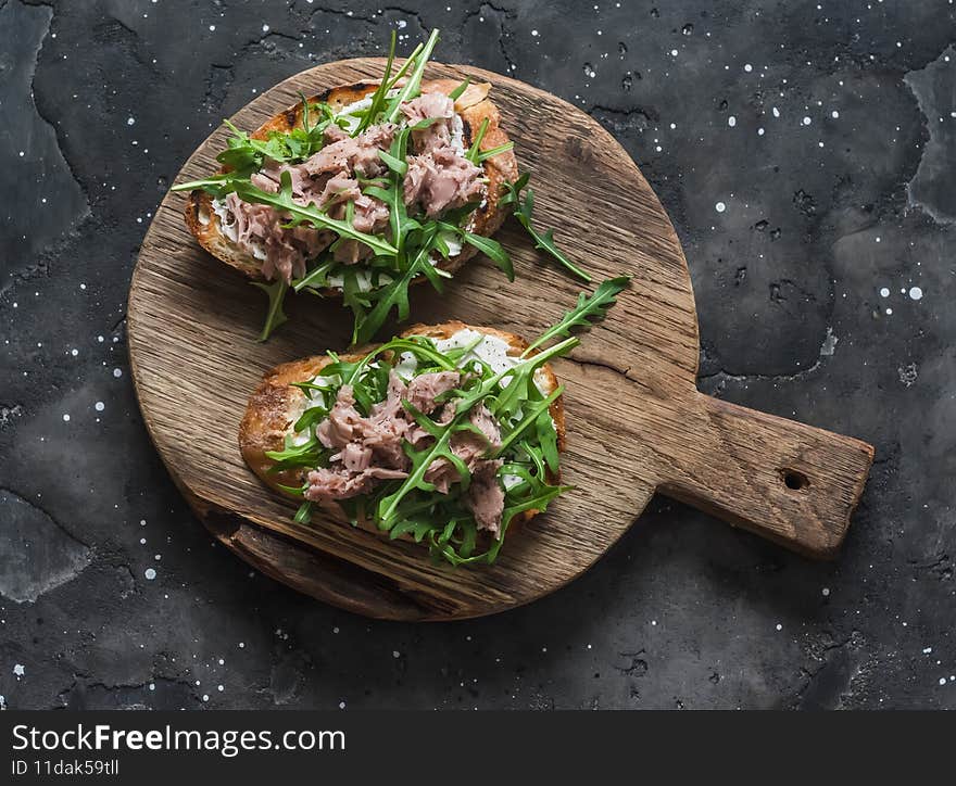 Snack sandwiches with canned tuna, cream cheese and arugula on a cutting board on a dark background, top view