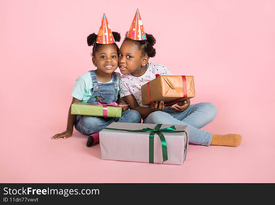 Holiday present, child& x27;s birthday concept. Two cute little excited african girls, wearing party hats, sitting on the