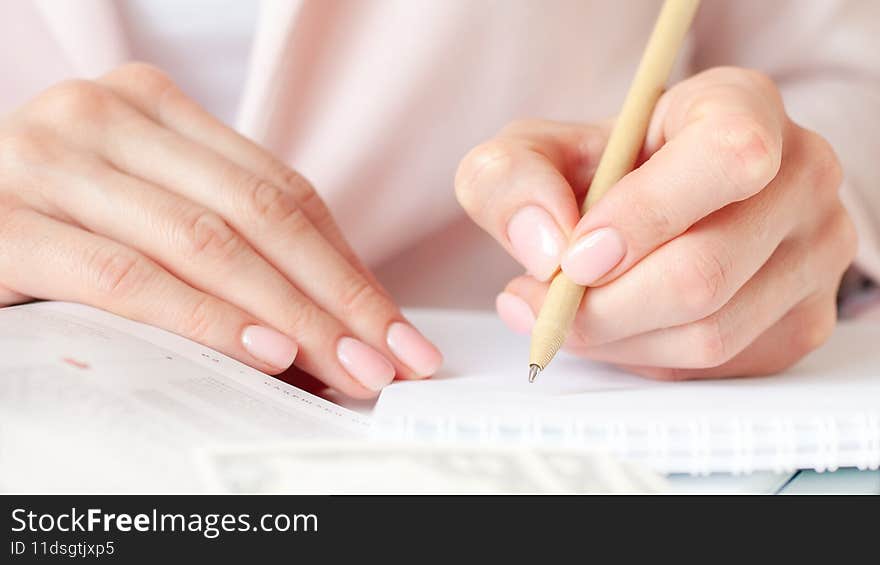 Closeup image of a woman writing down on a white blank notebook on wooden table. Pink background, selective focus. Business and education concept. Closeup image of a woman writing down on a white blank notebook on wooden table. Pink background, selective focus. Business and education concept