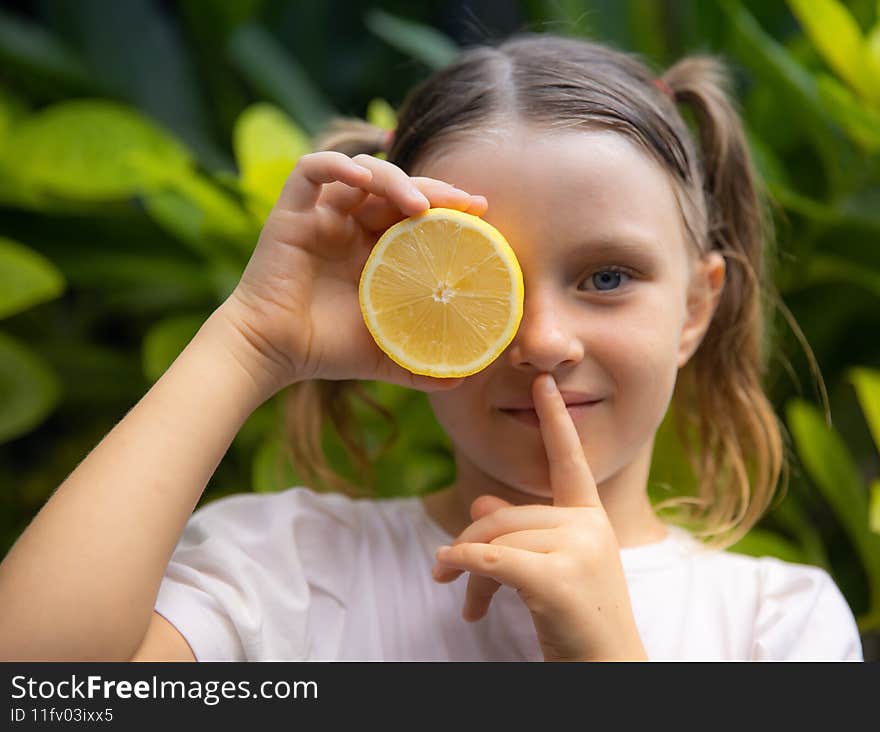 Close up portrait of little girl with lemon. Covering eye with half of fresh lemon. Girl puts finger on lips, making hush sign or quiet sound gesture. Selected focus on fruit. Bali, Indonesia