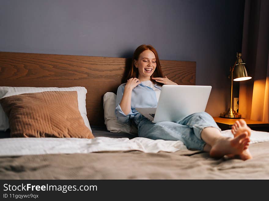 Joyful redhead young woman chatting online on notebook lying on bed at cozy bedroom.