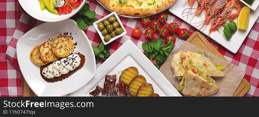 Traditional Italian cuisine concept flat lay. Sea food pizza, cooked shrimps, caramel ribs, ham pita sandwich, green olives and fresh cherry tomatoes over red plaid tablecloth