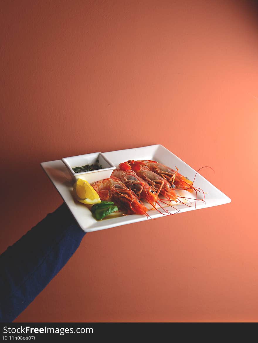 Cooked shrimps with lemon and sauce in a saucepan served on a white plate in hand over bright orange background. Sea food, healthy eating