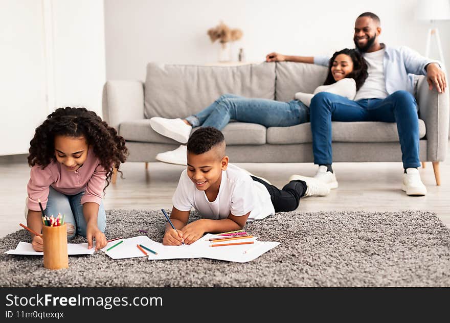 Family Weekend. Smiling African American kids drawing on paper with colorful crayons lying on the floor carpet in living room, happy parents relaxing and sitting on the couch in the blurred background
