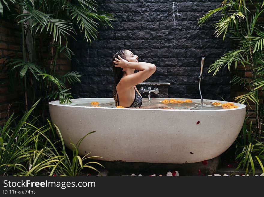 In the open-air bathroom near the brick wall with palm trees at the edges sits taking spa procedure girl while in the water float buds of flowers. High quality photo