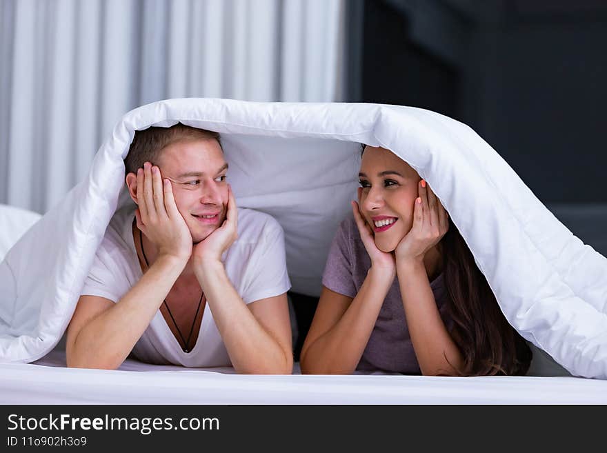 Portrait of young happy playful couple relaxing in comfortable cozy bed, cheerful smiling men and women having fun posing covering head with soft warm blanket duvet on sleepover
