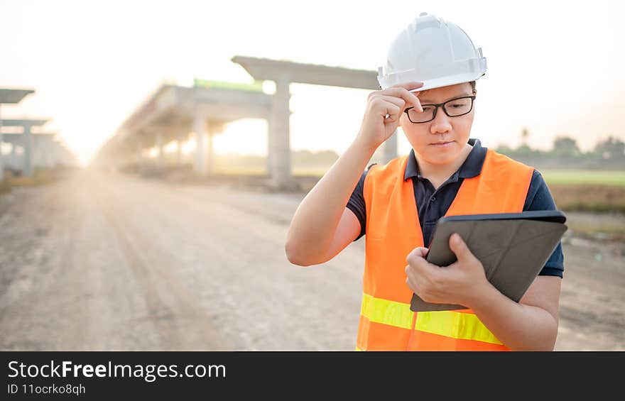 Smart Asian worker man or male civil engineer with protective safety helmet and reflective vest using digital tablet for project planning and checking architectural drawing at construction site. Smart Asian worker man or male civil engineer with protective safety helmet and reflective vest using digital tablet for project planning and checking architectural drawing at construction site