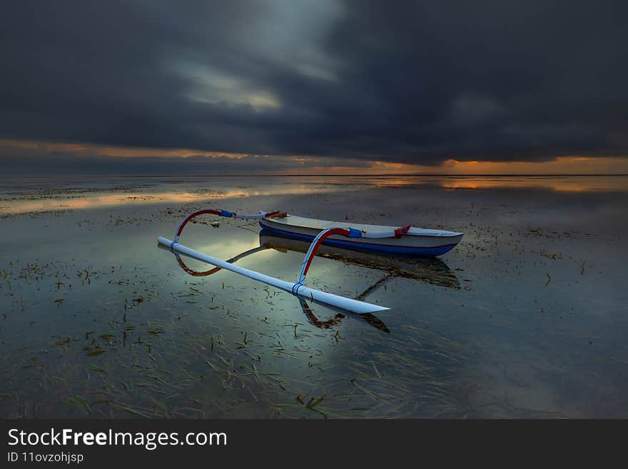 Seascape. Fisherman boat. Traditional Balinese boat jukung. Fishing boat at the beach during sunrise. Water reflection. Cloudy sky. Slow shutter speed. Soft focus. Sanur beach, Bali, Indonesia