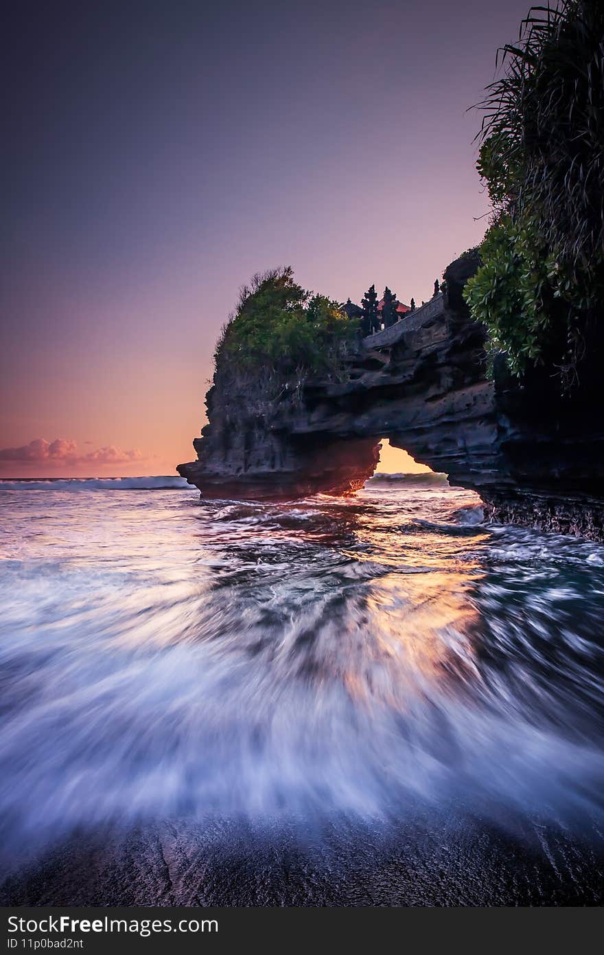 Natural arch. Batu Bolong temple on the rock during sunset. Seascape background. Motion milky waves on black sand beach. Copy space. Vertical layout. Tanah Lot, Bali, Indonesia