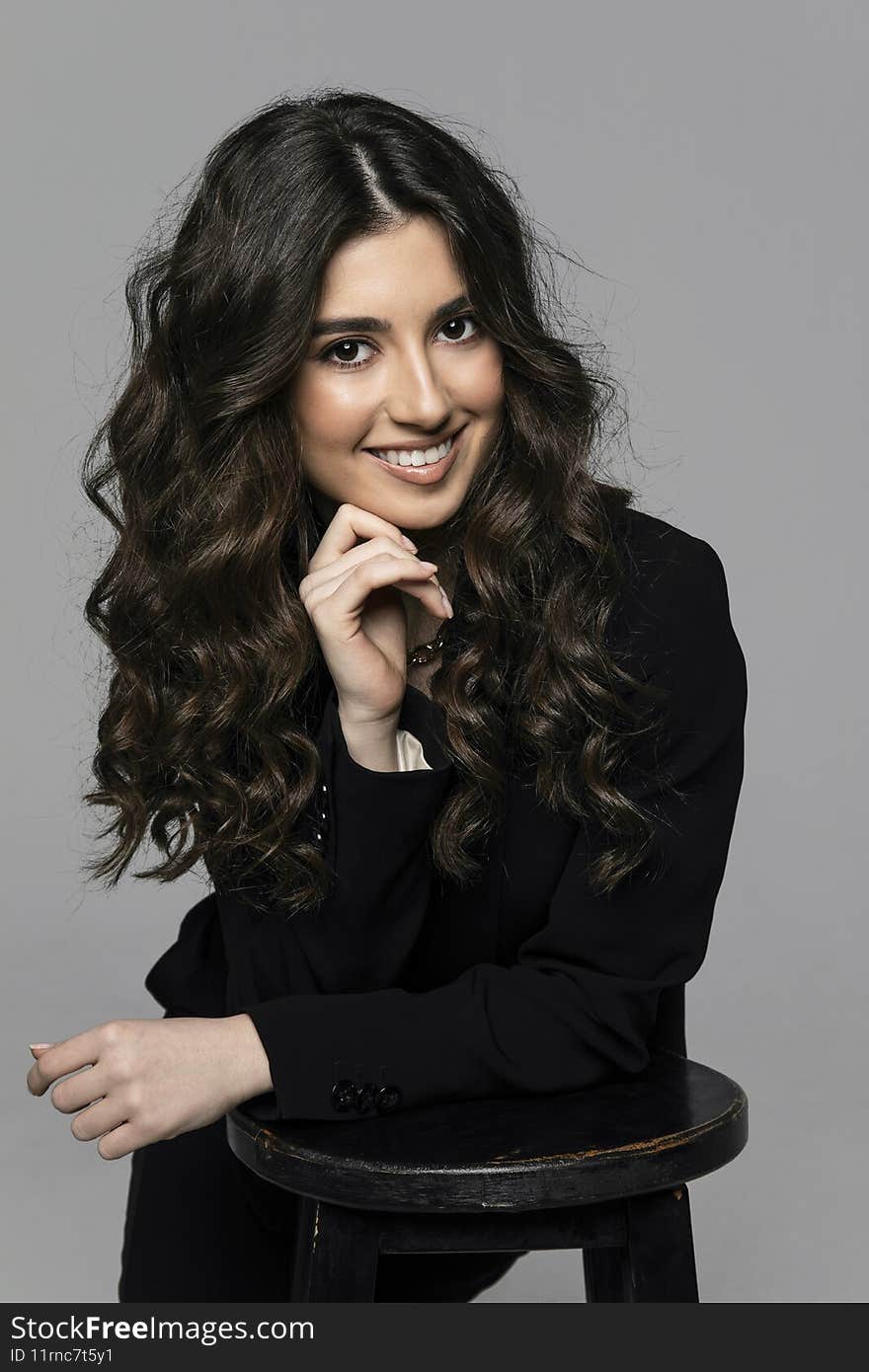 Beautiful sensual young woman curly hair with evening makeup wearing black suit, looking at camera, grey background.