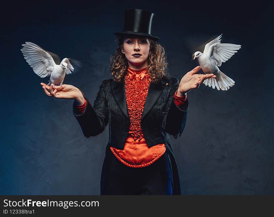 Portrait of a woman actor showing some tricks with white doves in dark background. Caucasian female artist wearing costume with top hat. Portrait of a woman actor showing some tricks with white doves in dark background. Caucasian female artist wearing costume with top hat.
